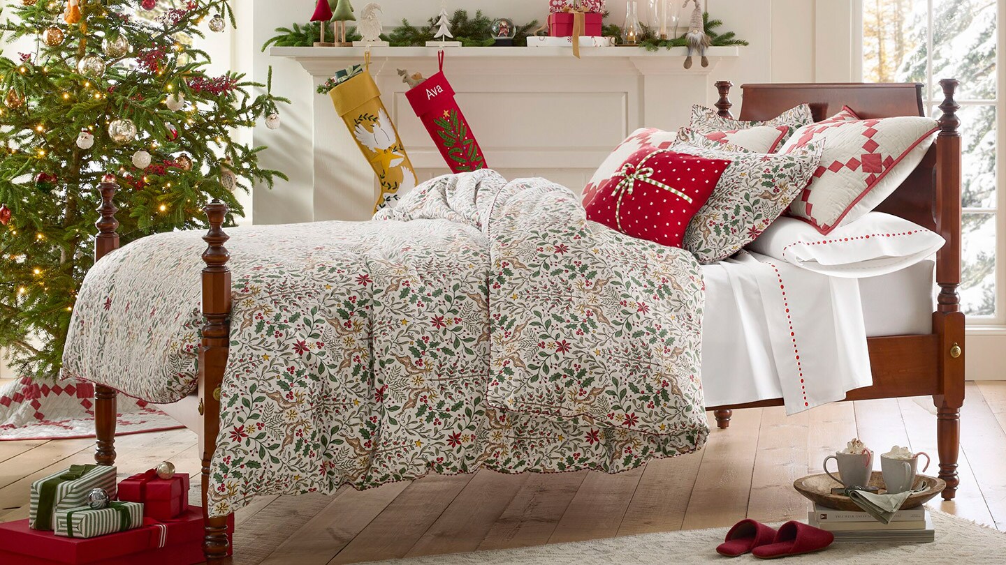 A bed with festive holiday bedding near a Christmas tree and stockings. Shop for bedding.